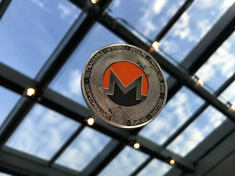 A silver Monero token with the orange XMR logo rests on a reflective glass surface beneath a skylight.
