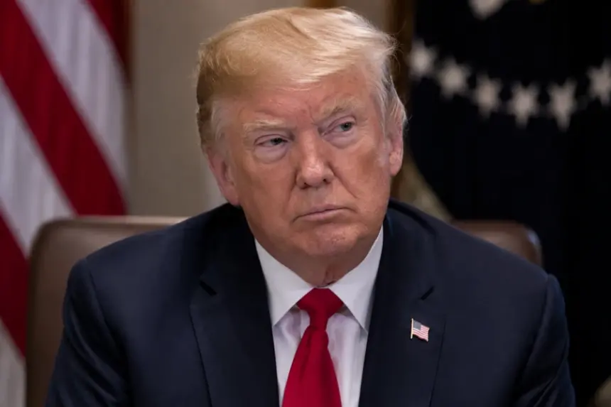 Donald Trump sits at a formal meeting table in a dark suit and red tie, with US flags blurred behind him.